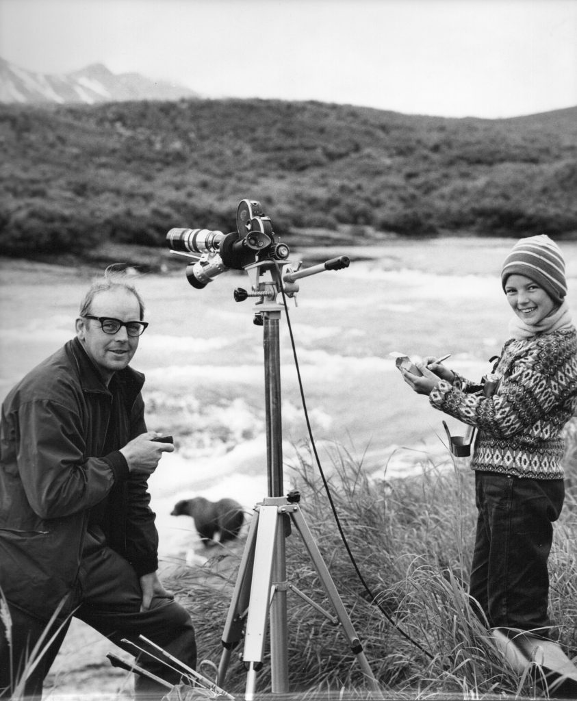 Svartvit bild på Sven och Pia, leendes mot kameran. 1961. En björn fiskar i bakgrunden.
